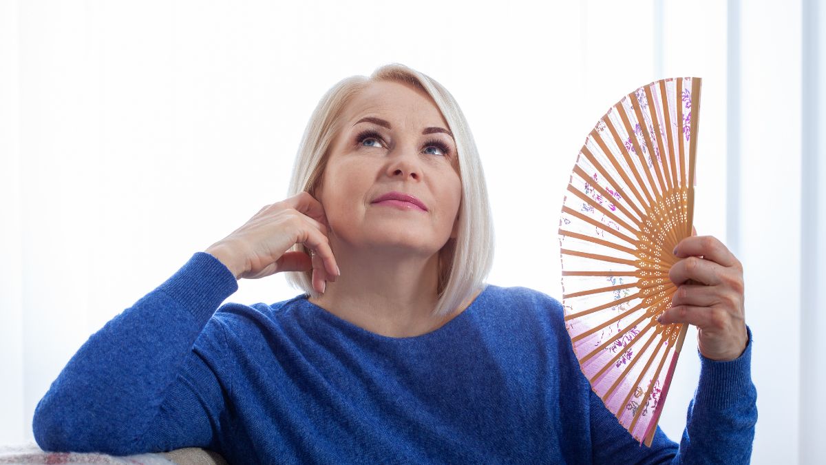 Egy középkorú nő gondolkodva ül, kezében egy ventilátorral, a perimenopauza tüneteire utalva.